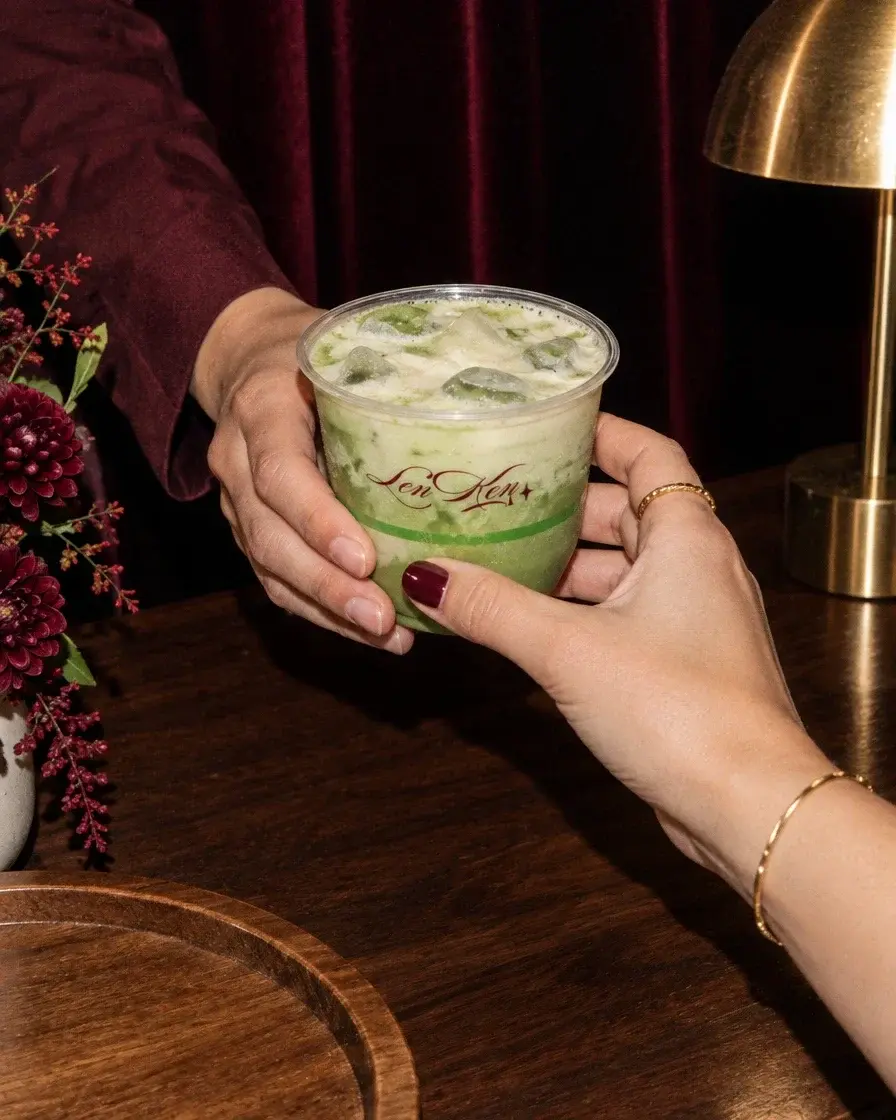 A Lenken matcha handed from one hand to another across the cart, beside red florals and a brass lamp.